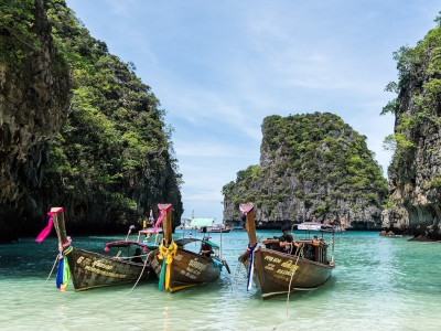 Typische thailändische Boote mit bunten Fahnen liegen am Strand von Phuket. Im Hintergrund sieht man einen mit üppigem Grün bewachsener Felsen